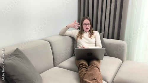 Video call chat continues as woman on sofa laughs with laptop at home. Female in glasses enjoys relaxed online conversation smiling during virtual talk. Lifestyle communication concept for