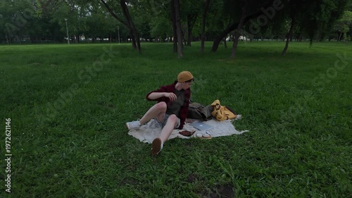Young woman stretching and packing laptop into backpack after working on blanket in green park during evening. End of remote work session in calm outdoor setting
