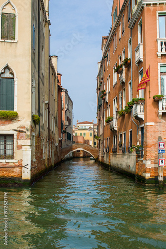 In Venice Italy a narrow canal slips between pastel palazzo walls and textured stone. Green water mirrors shutters balconies and trailing plants above aged masonry.
