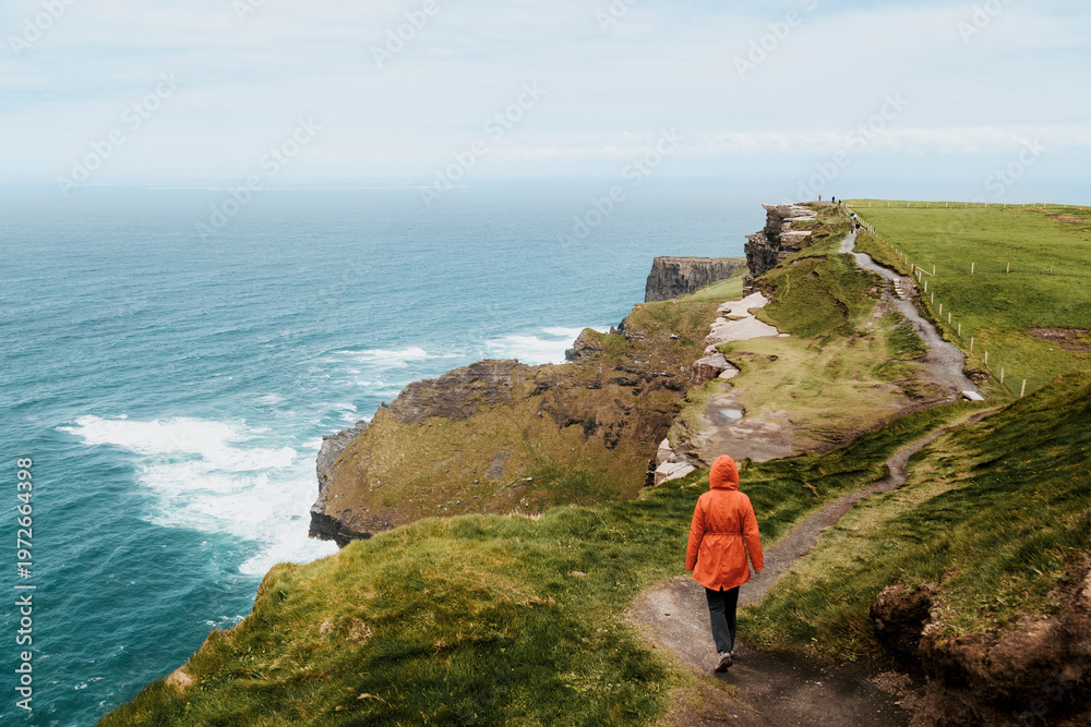Naklejka premium A person in a bright orange jacket walks along a narrow coastal path winding high above the churning blue waters of the Atlantic at the Cliffs of Moher.