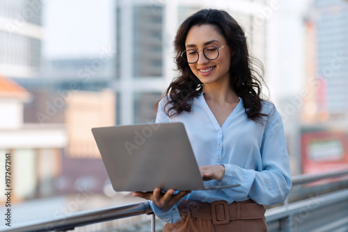 Free Download Cheerful businesswoman using her laptop outdoors in urban city area, lady leader being focused and happy as working - GFXLoad