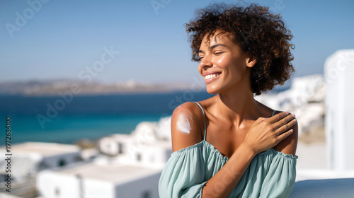 Happy smiling woman with curly hair applying sunscreen cream on shoulder, eyes closed, enjoying sun on Mediterranean terrace. Summer vacation, SPF skin protection, turquoise sea blurred background.