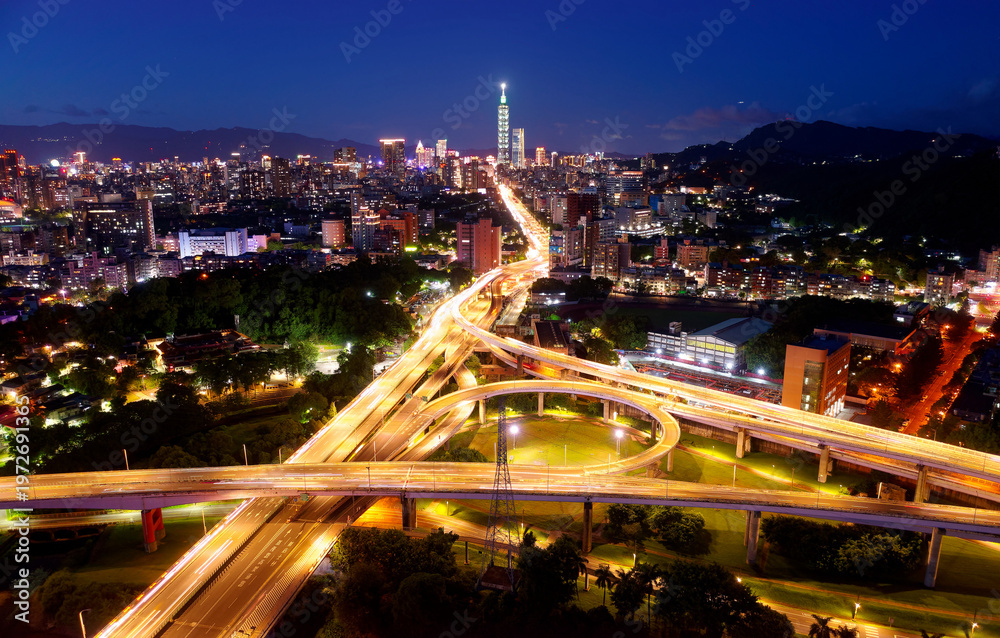 Obraz premium Aerial view over the circular interchange of Shuiyuan Expressway, Keelung Road & Fuhe Bridge in Taipei, with 101 Tower standing amid crowded buildings in Xinyi District & city lights dazzling at dusk