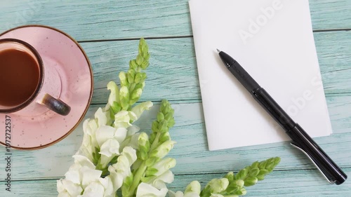 Cup of coffee sits beside a pen and notepad. White flowers bloom beside a rustic wooden surface. Pastel pink saucer adds delicate contrast to the scene. Black pen rests on clean, empty paper