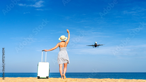 Woman waves goodbye from beachside, suitcase beside her
