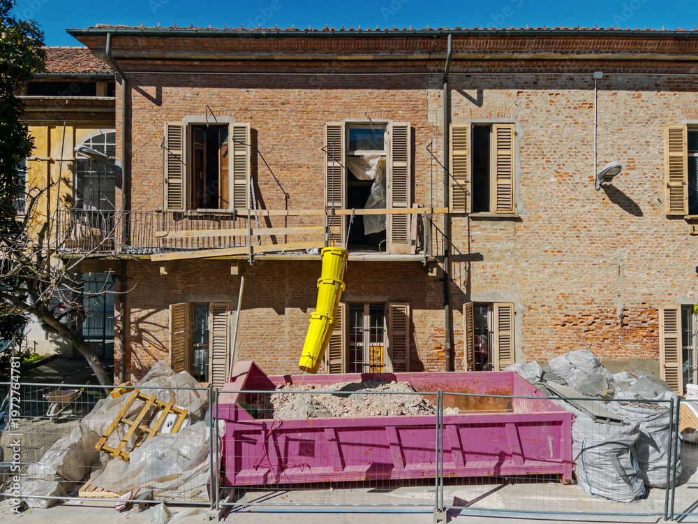 Fototapeta premium Construction site for the renovation of an old residential building. A yellow debris chute conveys the rubble into a roll-off container positioned at street level
