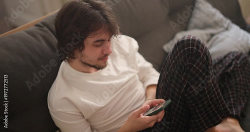 A young man in comfortable attire enjoys a quiet evening at home, focused on his smartphone and lost in thought