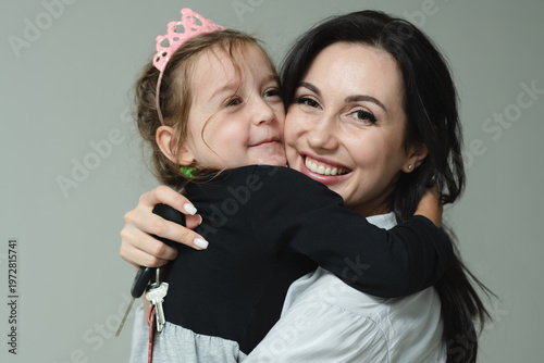 Mother and daughter is hugging and smiling. Mom hugs her little princess. Woman with keys in hand and little girl in pink crown embraces together. Facial expression. Selective focus