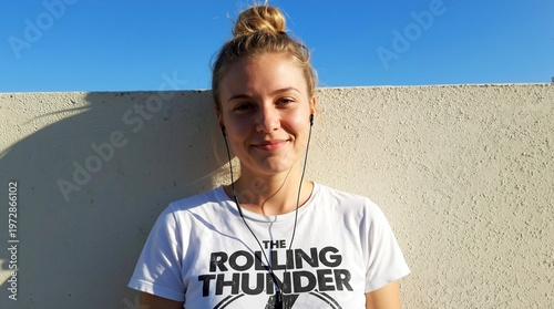 A portrait of a smiling young woman with blonde hair tied in a bun, wearing a white t-shirt and wired headphones, standing outdoors against a textured beige wall under a clear blue sky. Generative AI.