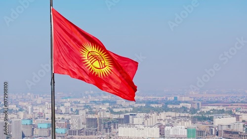 Red Kyrgyzstan Flag Waving Above Buildings of Bishkek City at sunny day with haze above the downtown.