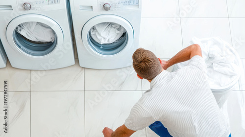 man crouches in front of two washing machines in a bright laundry room with a basket of white laundry beside him.