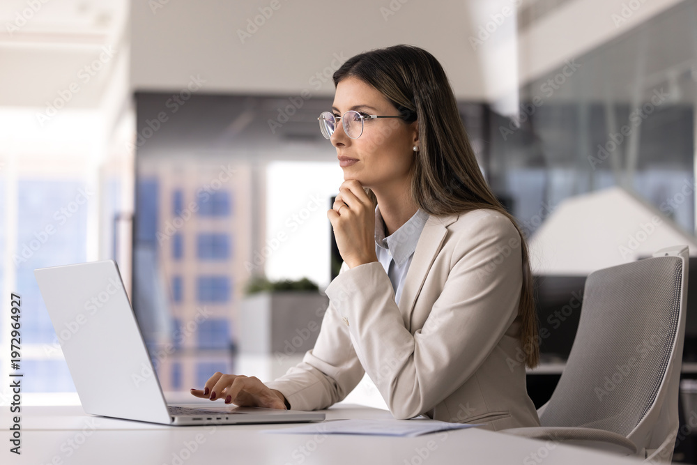 Fototapeta premium Confident young businesswoman in formal attire thoughtfully looks into distance while working on laptop seated at office desk. Decision making, strategy, and digital analysis in corporate workplace