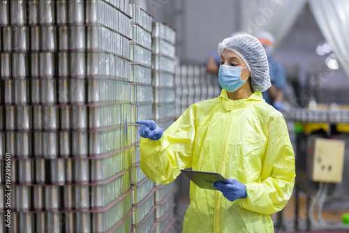 Quality control worker inspecting stacked canned fish products in seafood factory warehouse. Industrial food processing, packaging plant maintaining hygiene standards, export manufacturing compliance.