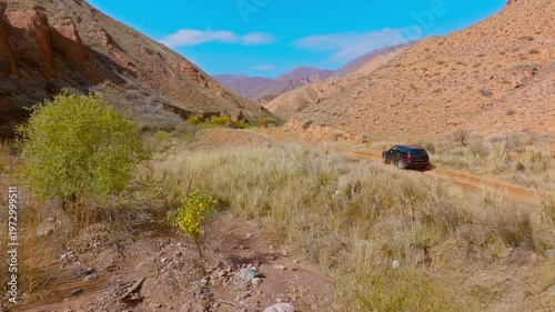 Black car navigates rugged dirt road through dry, mountainous canyon. Drone view with dolly forward movement.