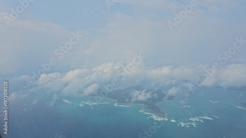 High-altitude aerial video overlooking a large, forest-covered Andaman island partially obscured by fluffy white clouds surrounded by the sea.