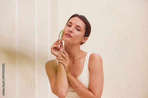 Woman holding a fresh aloe vera leaf near her cheek with both hands, gazing sideways with a serious calm expression. Ideal for aloe vera, dermatology, sensitive skin, or luxury clean beauty campaigns.