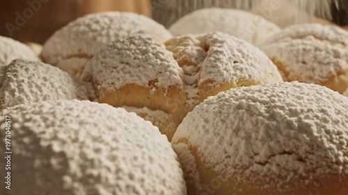 Close-up view of powdered sugar being sifted over freshly baked round sweet buns or brioche rolls resting on parchment paper and a wire cooling rack in a warm kitchen setting.