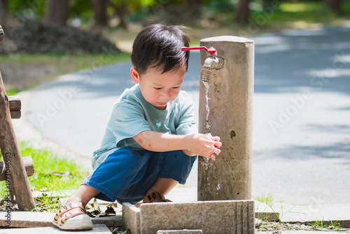 Young Asian boy in a light blue shirt crouching and washing his hands at an outdoor stone faucet. Blurred park background with natural light highlights a healthy and clean childhood lifestyle.