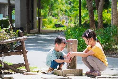 Happy Asian children, boy and girl, refreshing themselves with clean water from a park faucet on a hot summer day. Blurred green background highlights a cool and healthy childhood lifestyle.