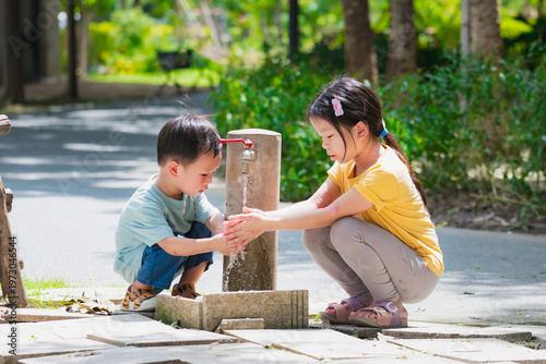Caring Asian older sister teaching her little brother how to wash hands at an outdoor park faucet. Blurred nature background highlights family bonding and healthy childhood hygiene habits.
