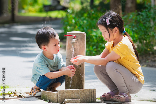 Two young Asian children, boy and girl, refreshing themselves and washing faces with clean water from a park faucet. Blurred nature background highlights healthy habits and summer joy.