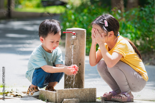 Two cute Asian children, boy and girl, splashing and washing faces with cool water from a park faucet. Blurred green background highlights refreshment and healthy childhood hygiene habits.