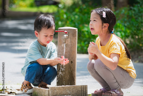 Candid portrait of Asian girl and boy refreshing themselves with cool water from a park faucet. Blurred green background highlights healthy childhood habits and summer joy.