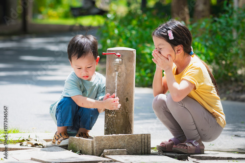 Two Asian children refreshing and washing faces with cool clean water from a park faucet. Blurred green background highlights healthy habits and summer joy.
