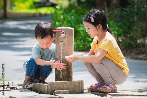 Asian boy and girl siblings washing hands and faces with clean water from a park faucet. Blurred nature background highlights healthy habits and summer refreshment.