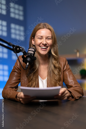 Portrait of smiling female host sitting at studio desk, holding script or notes papers while speaking into mounted broadcast microphone. Content creation for podcast, online show, or radio program