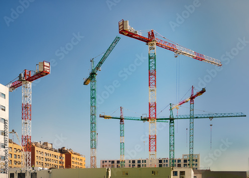 Multiple tower cranes at a construction site for new residential apartments under a clear blue sky, representing urban development, real estate investment, and the rising cost of housing.