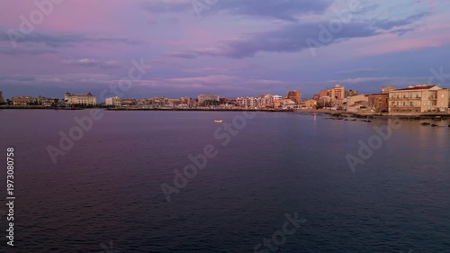 Dawn light illuminates Ortigia waterfront with pastel skies reflecting on calm waters, showcasing buildings and boats along the serene coastline