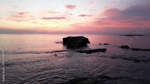 Serene coastal sunset over calm waters with a rocky outcrop silhouetted against vibrant pink and orange sky reflecting on the surface of the ocean
