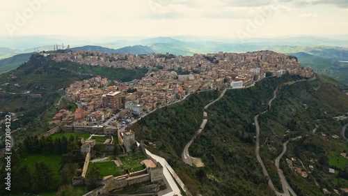 Aerial view of Enna, Sicily, showcasing the picturesque hillside town with winding roads, lush greenery, and historical architecture under a cloudy sky