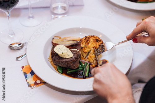 Close-up of a person cutting into a gourmet steak dinner with herb butter, potato gratin, and green beans in a red wine jus at a fine dining restaurant. Elegant atmosphere with red wine and cutlery.
