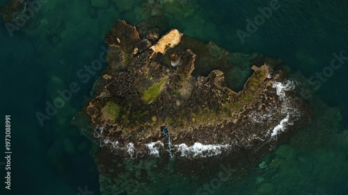 Aerial view of rocky island surrounded by turquoise waters, showcasing the gradual movement of waves crashing against the shoreline and revealing lush green vegetation