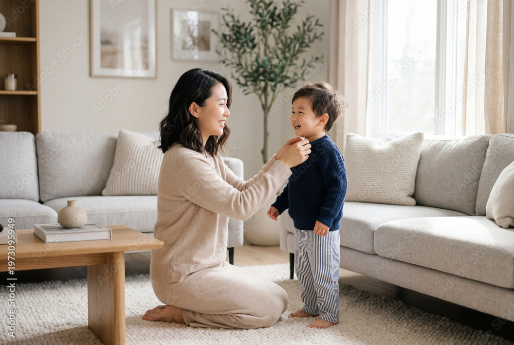 Fototapeta premium Loving asian mother carefully helping her young son put on his shirt in a bright living room