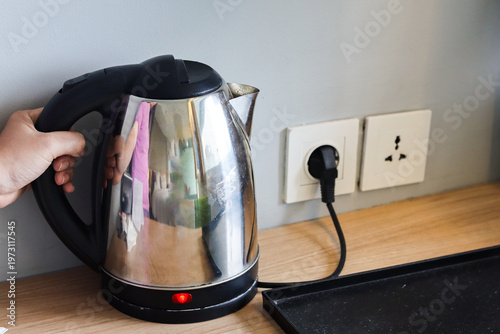 Electric kettle plugged into a wall outlet on a wooden table, with indicator light on, held by a woman's hand, used for boiling water in a kitchen or hotel room.