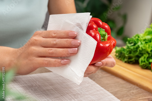 Close-up of hands gently wiping a vibrant red bell pepper with a paper towel. The scene suggests careful food preparation in a bright kitchen. Fresh green lettuce sits nearby on a wooden surface
