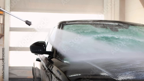 High pressure water jet spraying foam on black car windshield at professional car wash station, power washing vehicle front glass, auto detailing cleaning service indoors
