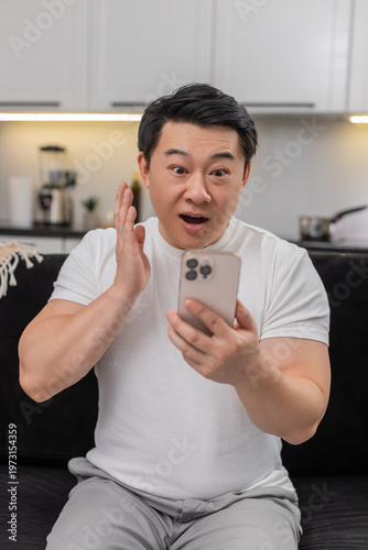 Asian man at home reads good news on smartphone says wow eyes wide smiles and starts typing quick reply. Chinese guy on sofa shares update in chat sends thanks adds heart emoji feels relief and hope