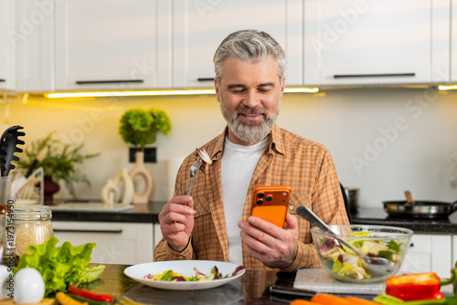 Middle-aged man enjoying salad while checking smartphone for calorie tracking weight loss plans in kitchen indoors. Mature guy smiling after healthy diet choice supporting fitness nutrition at home.