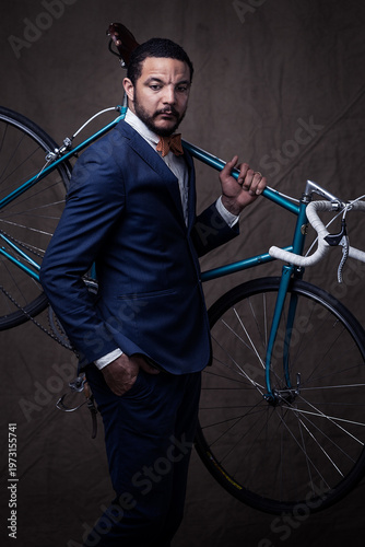 Dapper man in a navy blue suit and orange bow tie carrying a vintage teal bicycle on his shoulder. Elegant studio portrait with a classic backdrop, blending urban cycling style with formal fashion.