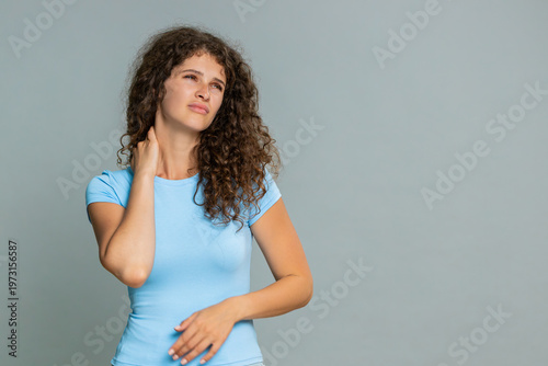 Woman experiencing neck pain rubbing tense muscles with tired, uncomfortable and exhausted facial expression. Girl on gray background demonstrates discomfort stress symptoms after working long hours.