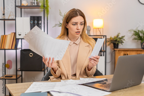 Middle-aged businesswoman at home office looks at bills invoices receipt on laptop sees costs go up frowns confused and unhappy. Freelancer girl at table writes savings plan sets targets prints page