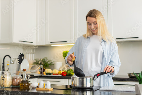 Middle-aged woman stirs hot soup with ladle on electric cooktop adding salt and pepper for healthy lunch at home kitchen. Young girl watches a cooking lesson learning nutrition habit for family meal.