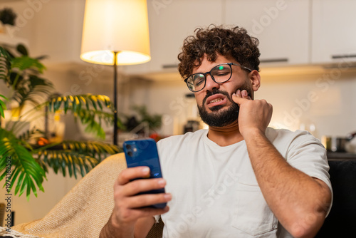 Εκτύπωση καμβά Arabian man at home feels tooth pain holds cheek opens smartphone search for dentist finds time