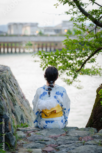 Fotografie Rear view of a woman in a blue and white floral yukata sitting on a rock by the Uji River looking towards a bridge