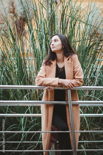 Portrait of a beautiful dark-haired woman in a beige coat standing among green plants. Vertical photograph.