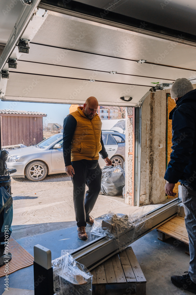 Fototapeta premium Man in yellow vest inspects delivery items inside a garage with an open door, revealing a car parked outside and a sunny day visible